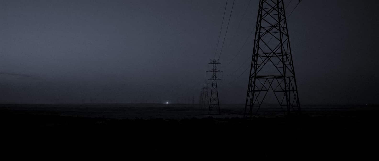 A line of high-voltage transmission towers receding to the horizon at blue hour, with a single light at a distant substation